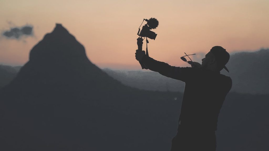 Silhouette of a man filming with camera at sunset with mountain in background.